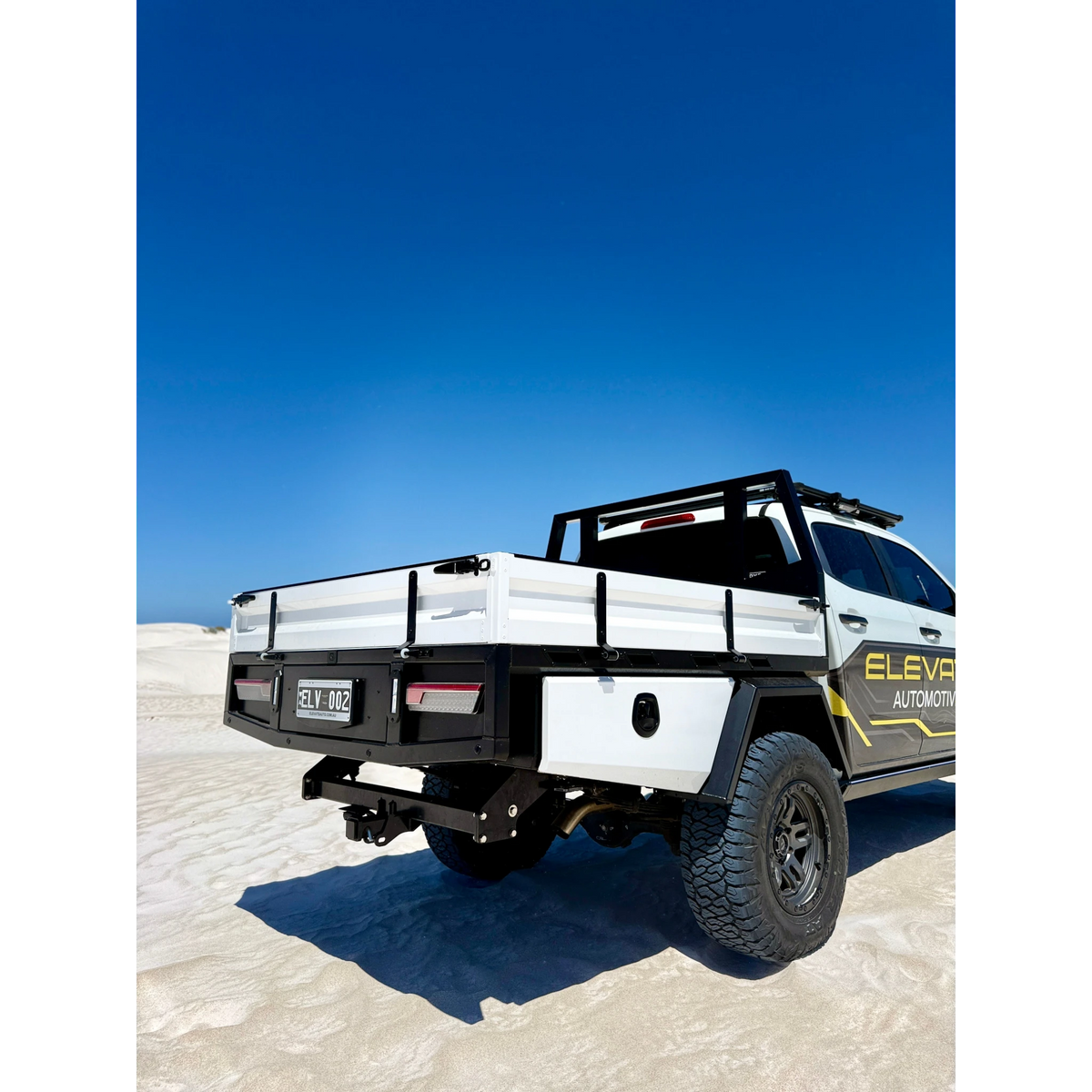 Rear three-quarter view of the aluminium tray fitted to an Elevate Automotive ute on white sand, showing the drop sides with their tie rails, the rear bar with recessed lights and number plate, the tow hitch and the underbody toolbox