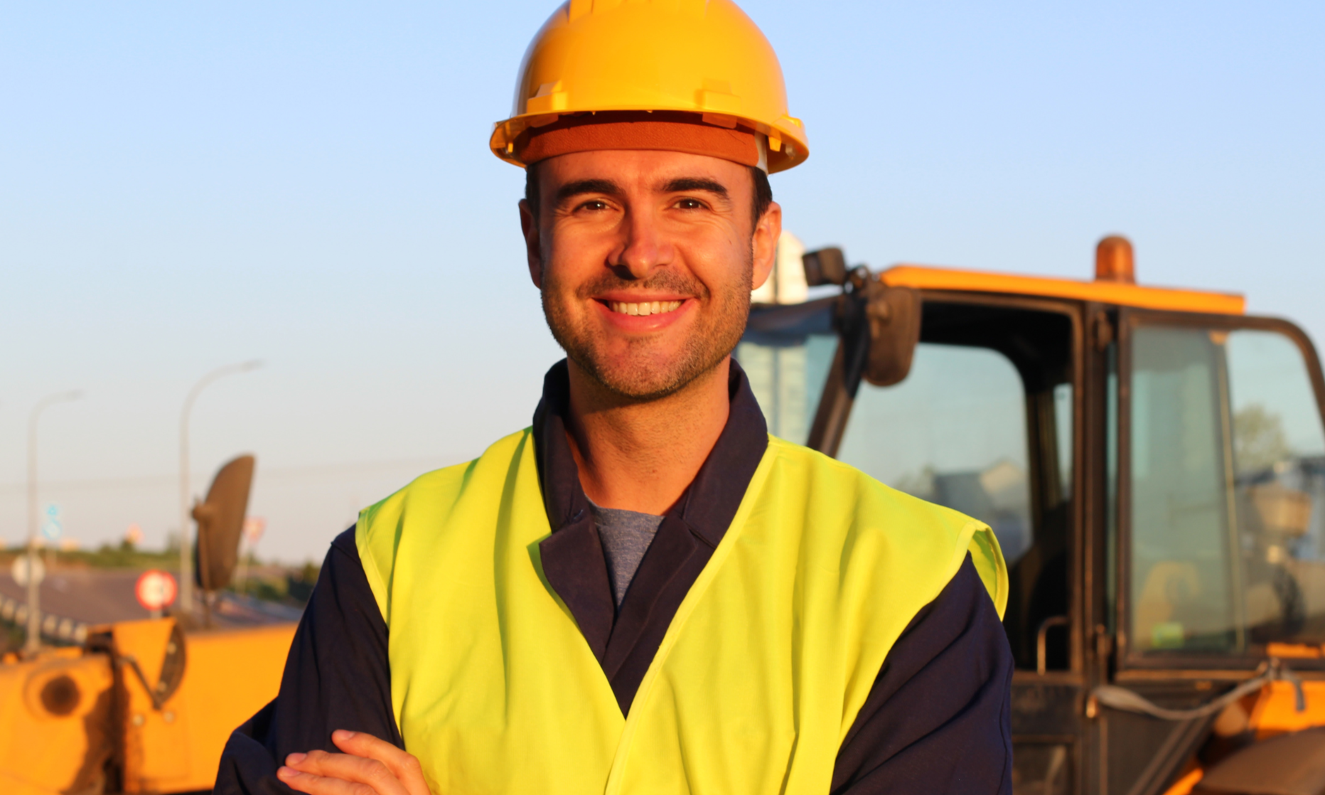Man wearing a yellow hard hat and safety vest standing in front of construction equipment.