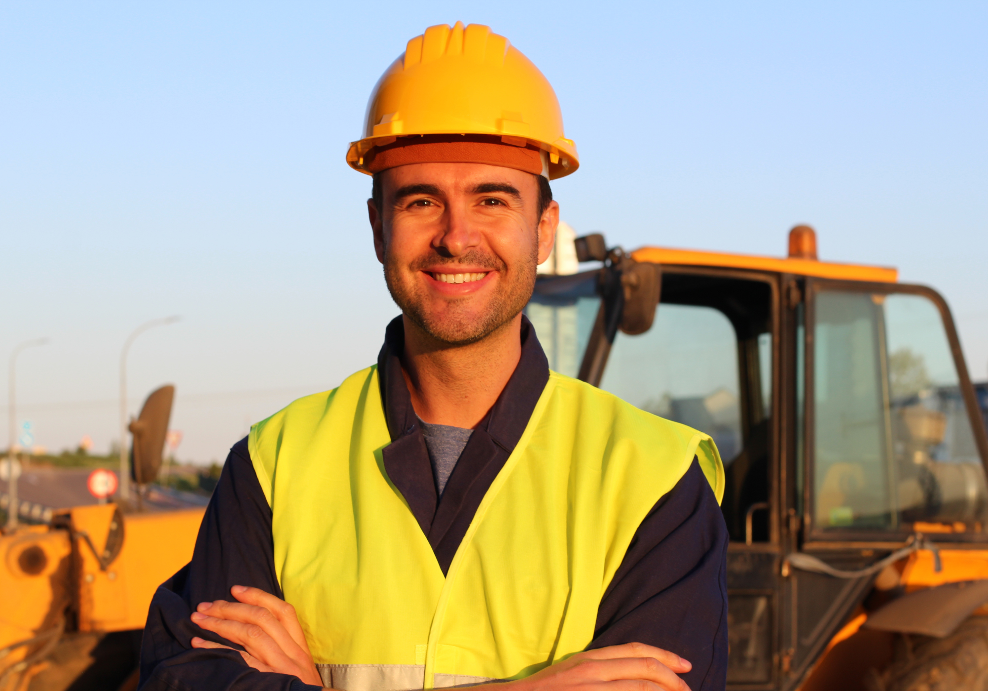 Man wearing a yellow hard hat and safety vest standing in front of construction equipment.