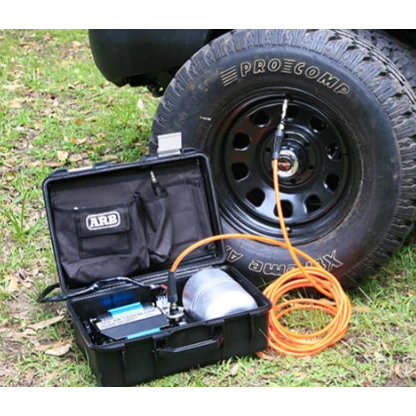 ARB twin piston compressor in its open travel case beside a vehicle on grass, the orange air hose connected to a Pro Comp wheel on a mud terrain tyre