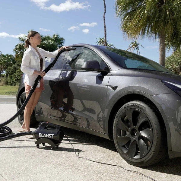 Woman using a Blahst Air Maxx Dryer on a gray electric vehicle with palm trees in the background.