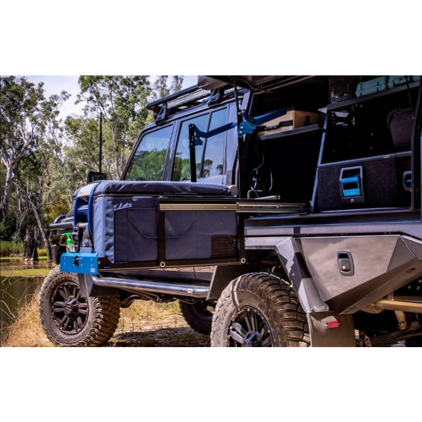 A Toyota LandCruiser troop carrier parked by water with the rear open, a fridge in a padded cover sitting out on the MSA drop slide and a canvas awning arm folded above it