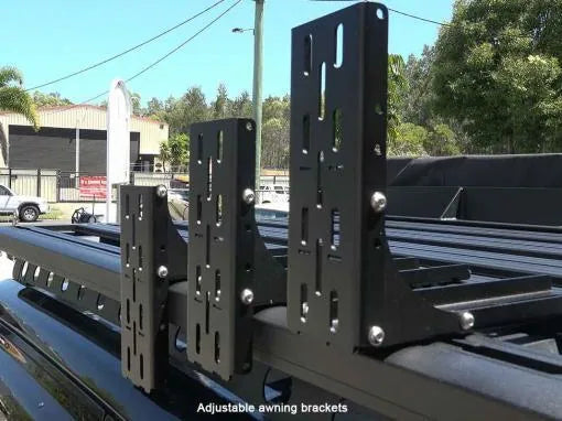 Adjustable awning brackets on a vehicle roof with a blurred background of trees and buildings.