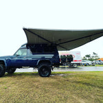 Blue SUV with a large awning extended outdoors on a grassy area.
