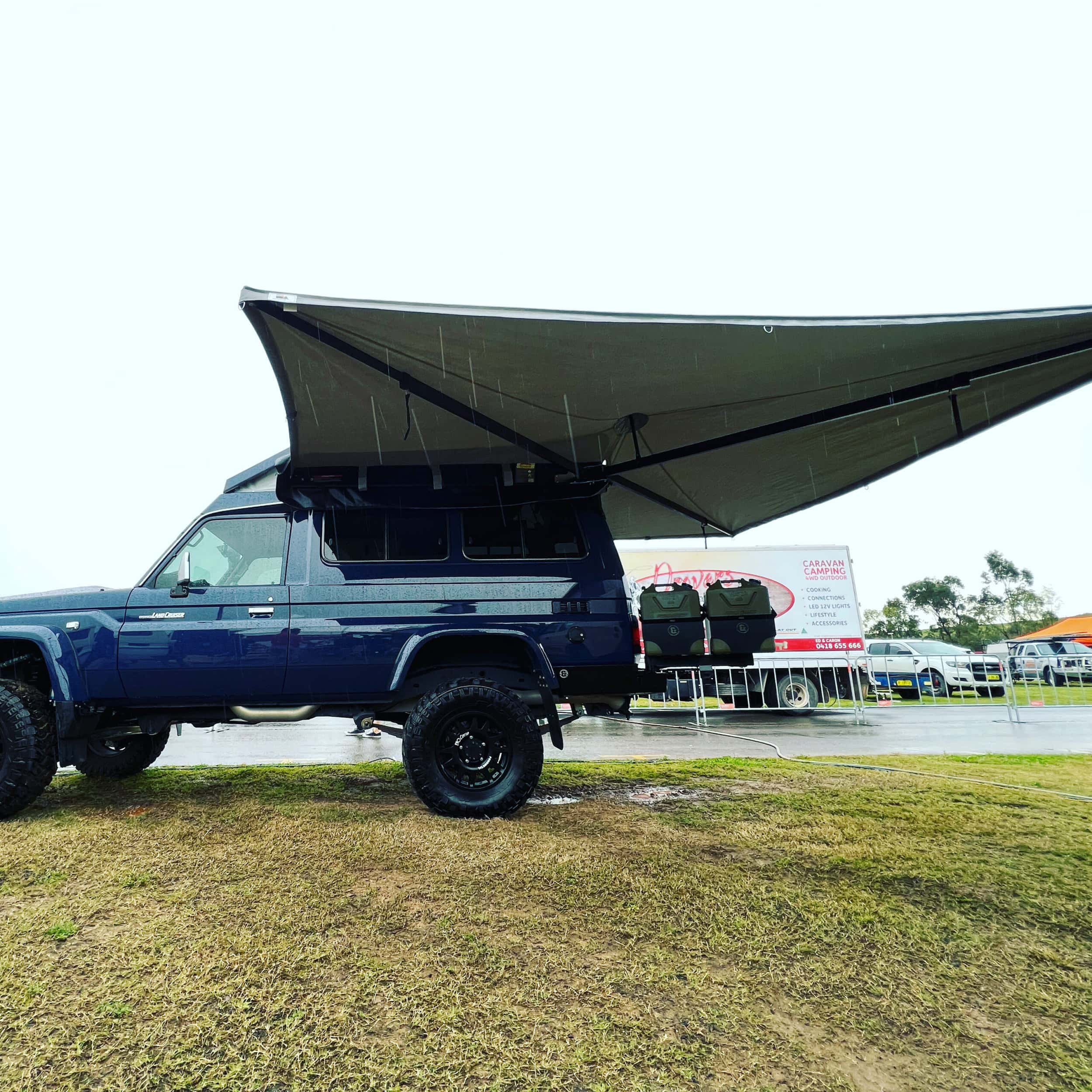 Blue SUV with a large awning extended outdoors on a grassy area.