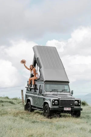 Person sitting on a Land Rover Defender with an open roof in a grassy field.