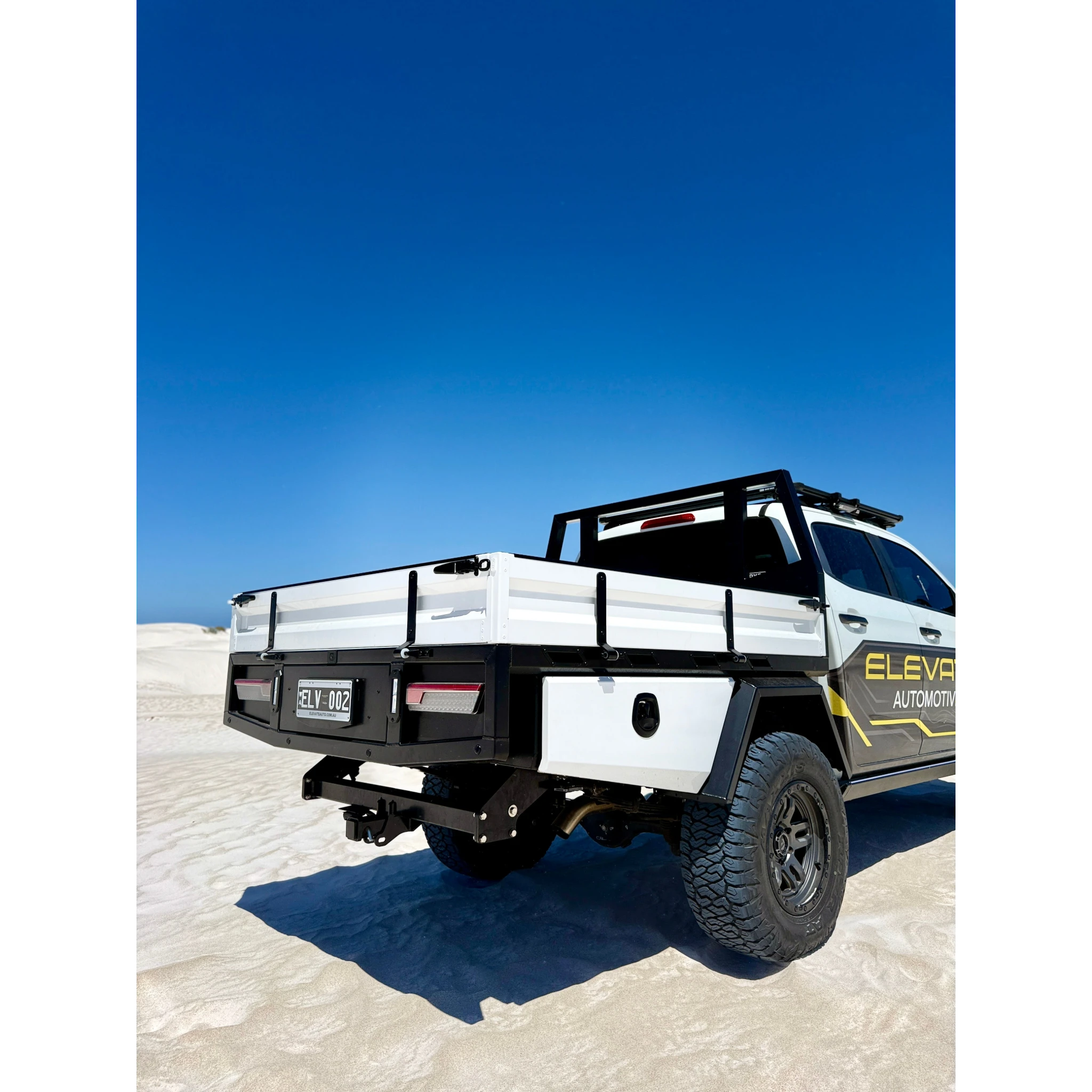 Rear three-quarter view of the aluminium tray fitted to an Elevate Automotive ute on white sand, showing the drop sides with their tie rails, the rear bar with recessed lights and number plate, the tow hitch and the underbody toolbox