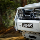 Close-up of a white Toyota vehicle's front bumper with LED lights and a blurred natural background.