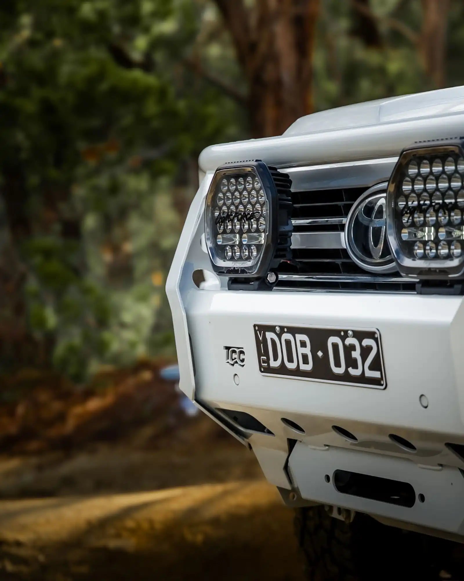 Close-up of a white Toyota vehicle's front bumper with LED lights and a blurred natural background.