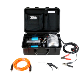 ARB twin piston compressor in its black travel case with the lid open, the twin blue cylinders and polished air tank inside, with the orange hose, blower gun and battery clamp leads laid out in front