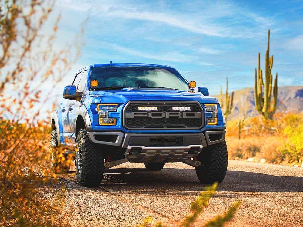 Blue F150 raptor truck on a desert road with cacti and mountains in the background with Lazer Lamps Grille Kit – Ford F-150 Raptor (2017–2020)