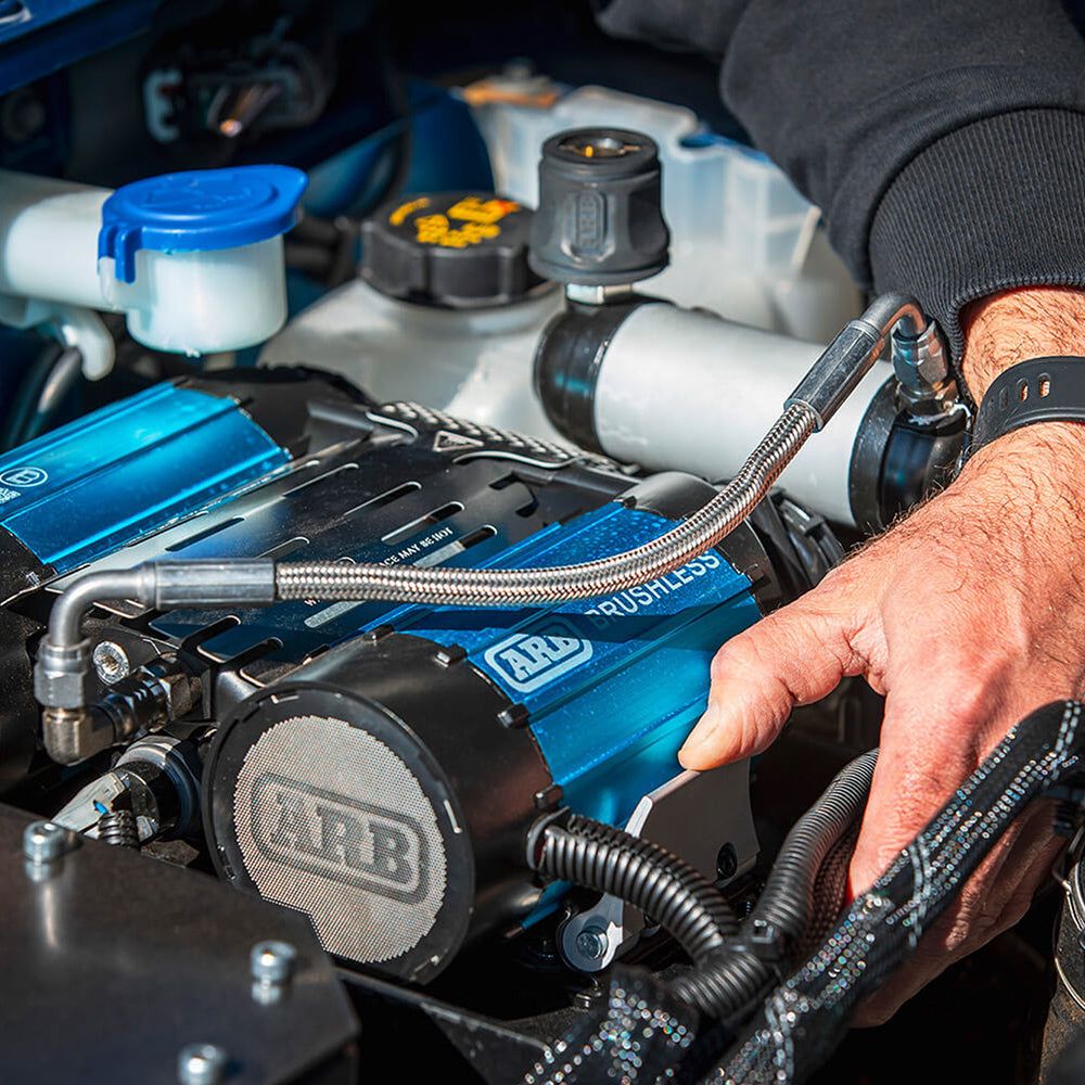 ARB brushless twin compressor being fitted in an engine bay, the blue anodised cylinders and braided air line in place under the bonnet with a hand resting on the housing