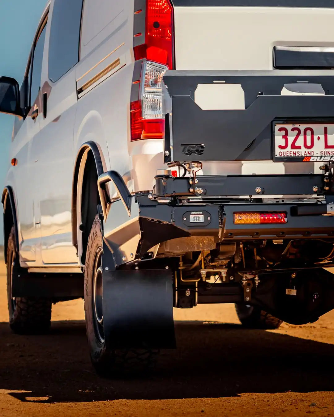 Close-up of a white van's rear with a black bumper on a dirt road.