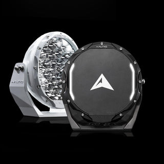 A white and a black ALTIQ Rogue 8.5 driving light photographed against black, the white one showing its reflector face and the black one its arrow-marked cover with the illuminated position-light strip curving across it