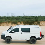 White van modified for off-road use on a sandy terrain with greenery in the background