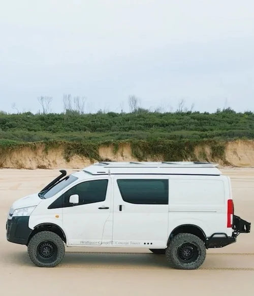 White van modified for off-road use on a sandy terrain with greenery in the background