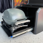 Portable grill with a hose attached inside an open toolbox on a concrete surface.