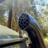Close-up of a car exhaust pipe with a honeycomb design, set against a natural background of trees.