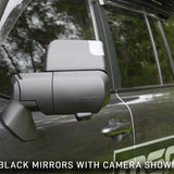 Car side mirror with built-in camera on a vehicle, surrounded by greenery.