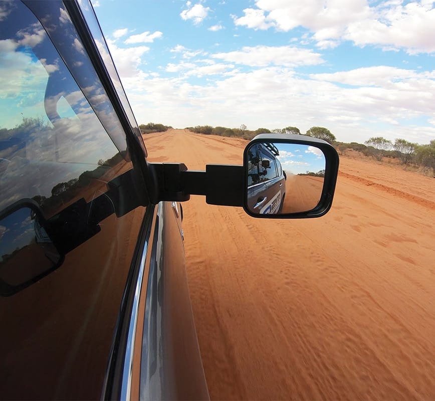 Side view mirror of a vehicle on a desert road with blue sky and clouds.