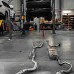 Exhaust system on the floor of a garage with a vehicle on a lift in the background.