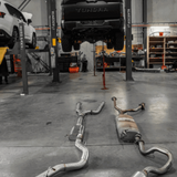 Exhaust system on the floor of a garage with a vehicle on a lift in the background.