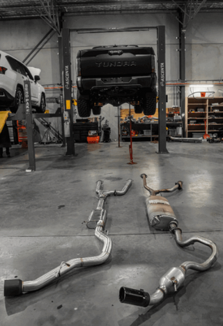 Exhaust system on the floor of a garage with a vehicle on a lift in the background.