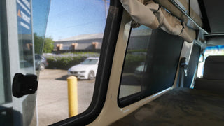 Interior view of a vehicle with open window and curtains, showing a parking lot outside.
