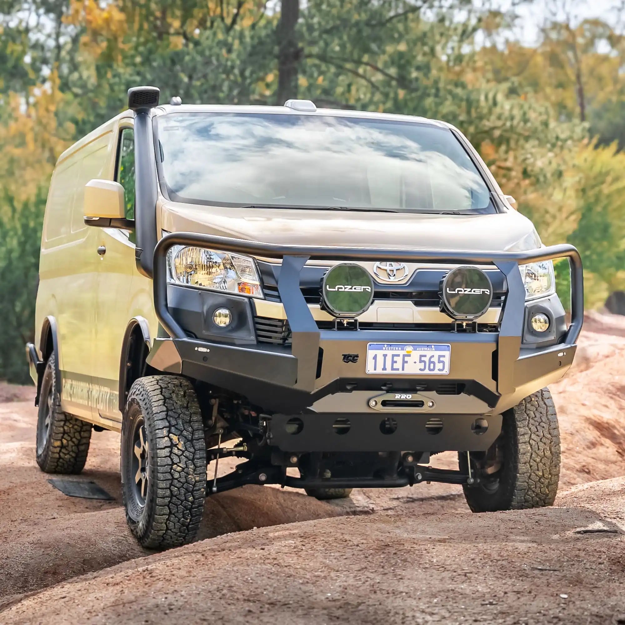 Gold van with a large front bumper on a dirt road with trees in the background