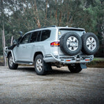 Silver SUV with spare tires on a gravel road surrounded by trees
