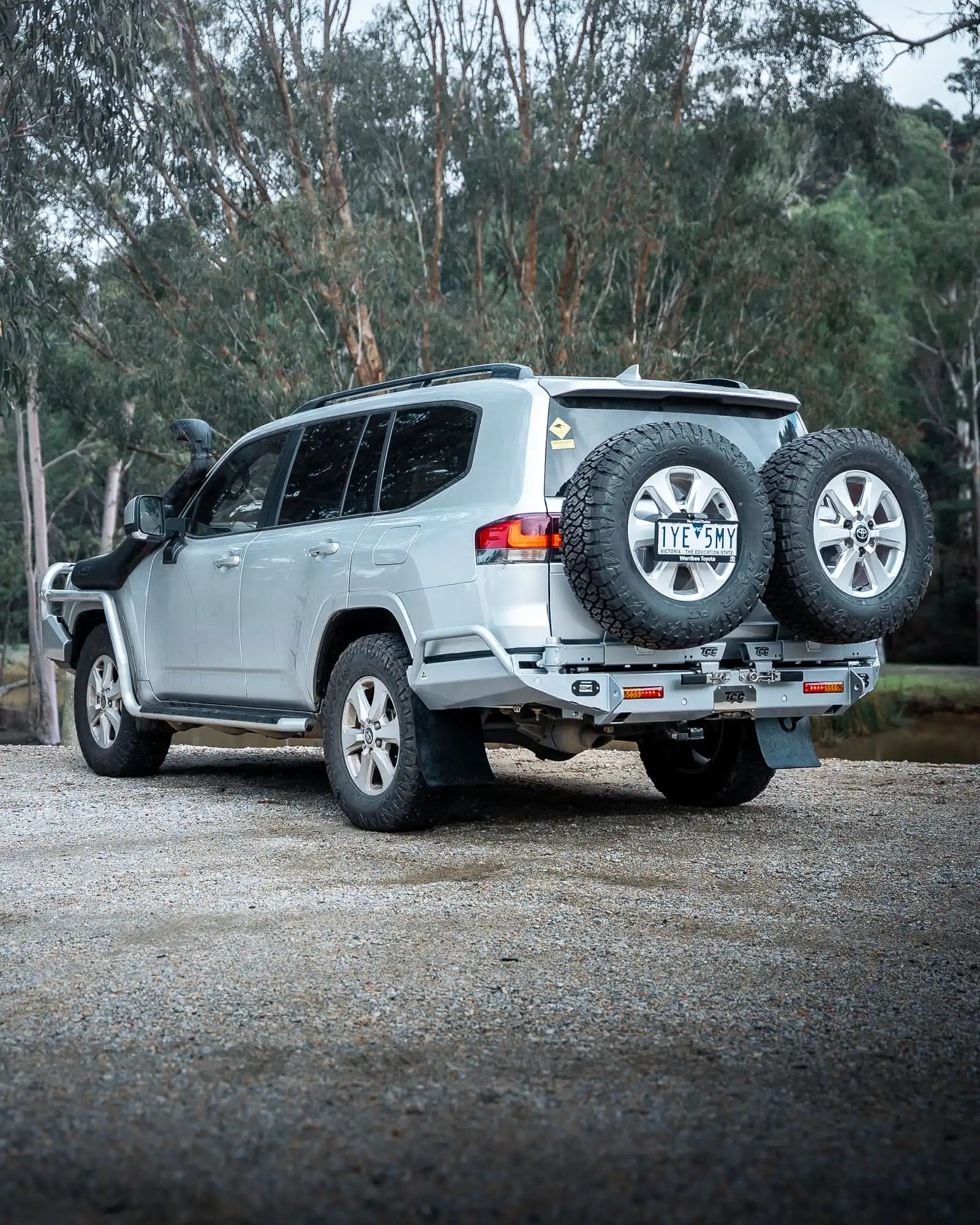 Silver SUV with spare tires on a gravel road surrounded by trees