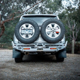 Back view of a silver SUV with spare tires on a gravel road surrounded by trees.
