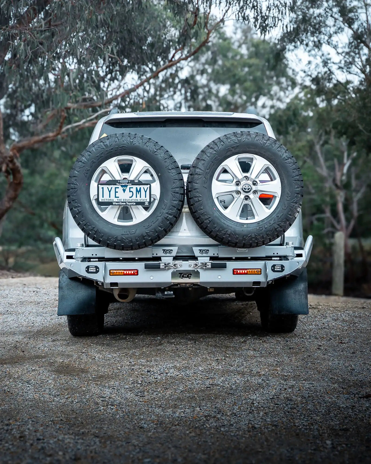 Back view of a silver SUV with spare tires on a gravel road surrounded by trees.