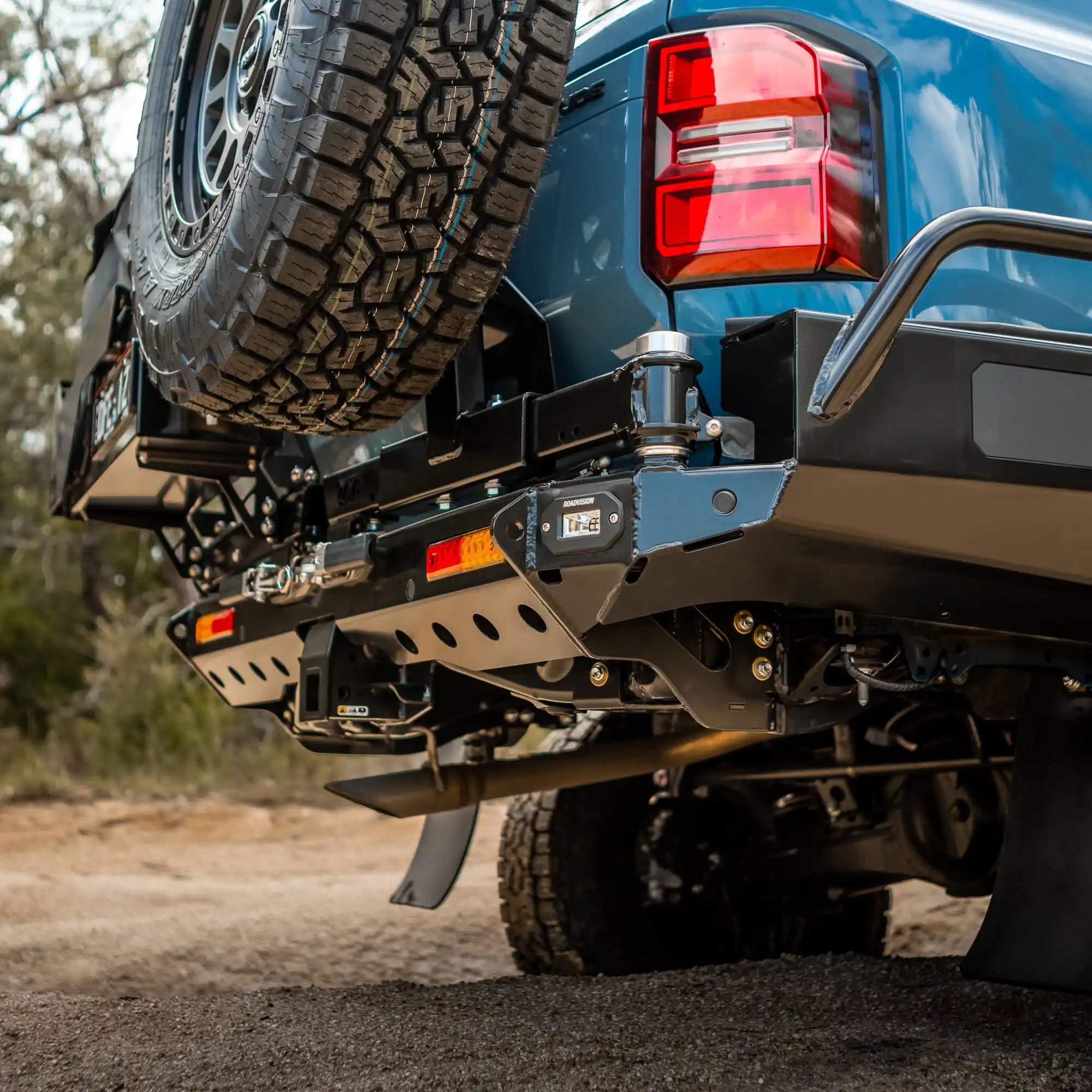 Close-up of a blue truck's rear with tire and bumper on a dirt road.
