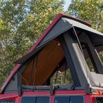 Red vehicle with an open roof top tent against a tree-lined background