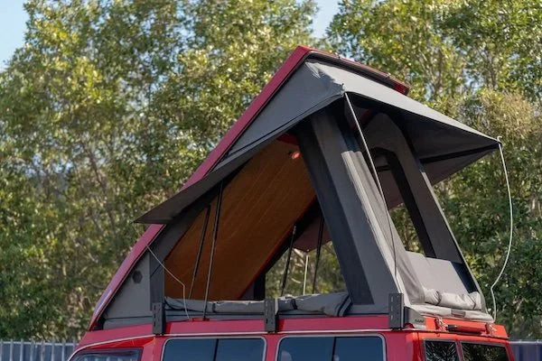 Red vehicle with an open roof top tent against a tree-lined background