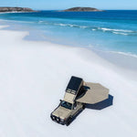 Beige off-road vehicle with a roof rack on a sandy beach with blue ocean and sky.