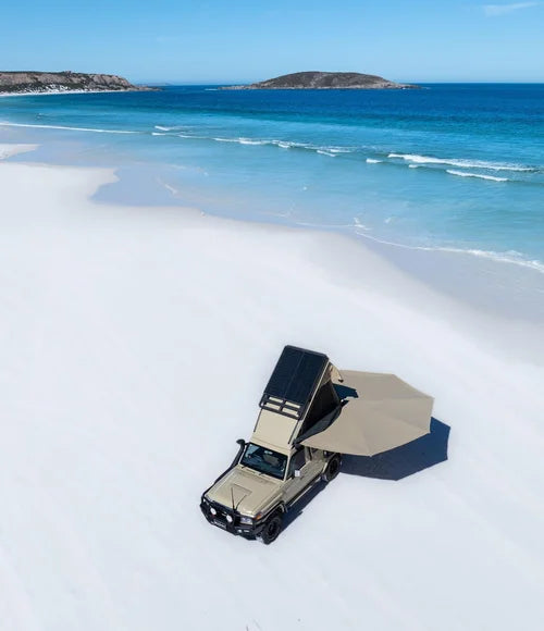 Beige off-road vehicle with a roof rack on a sandy beach with blue ocean and sky.