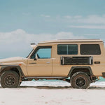 Beige off-road vehicle with a camper shell on a sandy beach with ocean and sky in the background