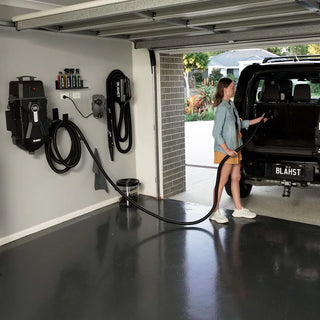 Woman using a car vacuum system in a garage with various cleaning tools on the wall.