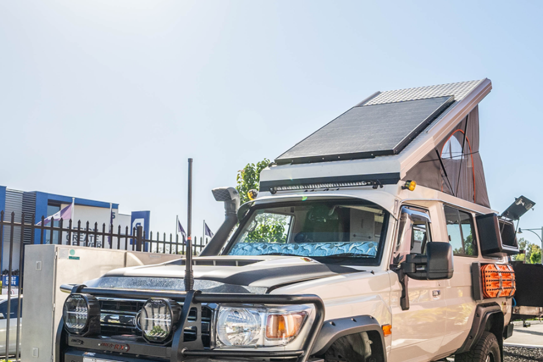 Beige off-road vehicle with a rooftop tent and solar panel on a clear day.
