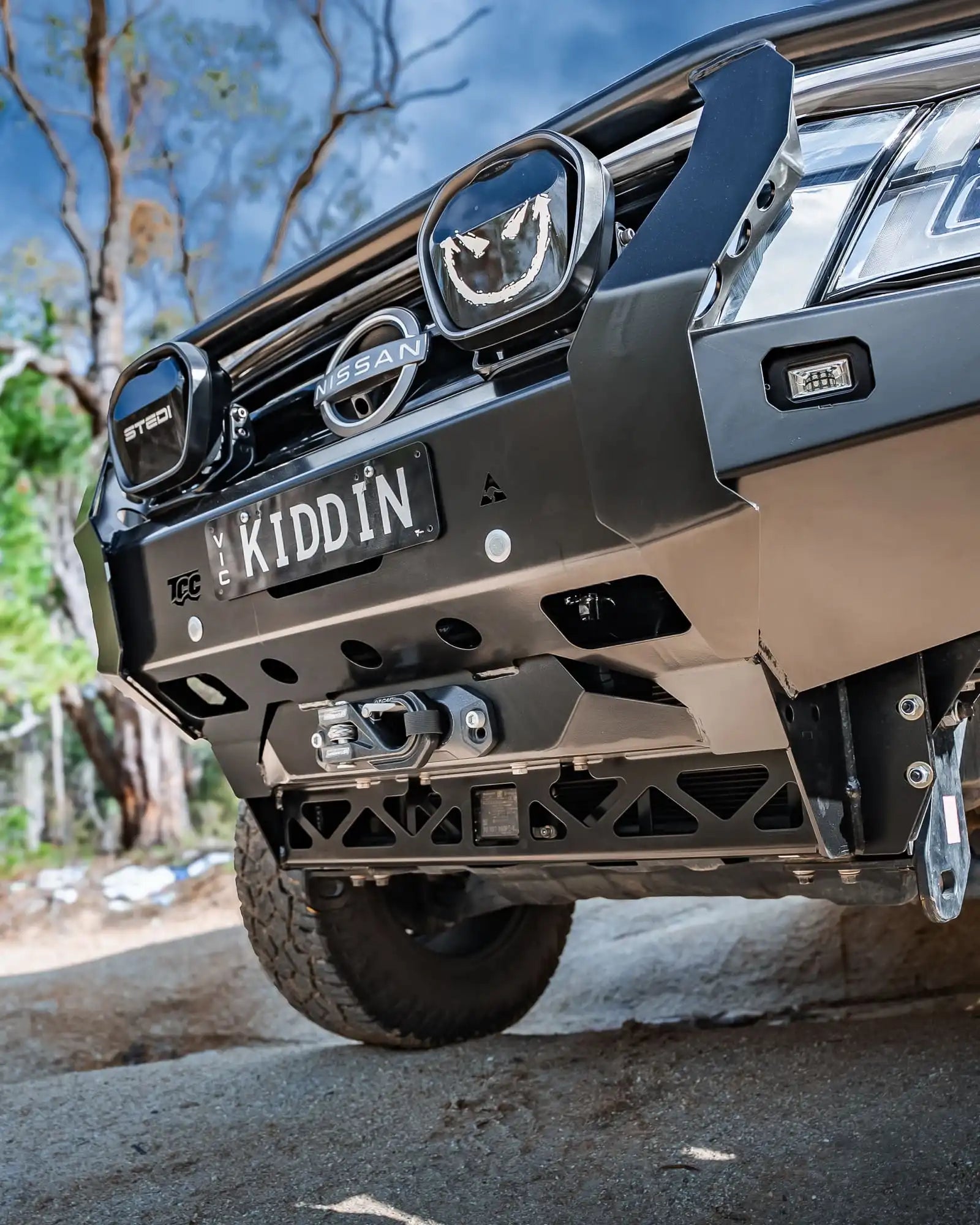 Close-up of a Nissan vehicle's front bumper with 'KIDDIN' license plate in an outdoor setting.