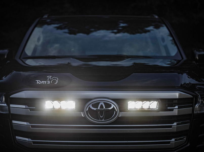 Close-up of a Toyota vehicle's front grille with illuminated headlights and Lazer Lamps Grille Kit – Toyota Land Cruiser 300 Series (2022+)