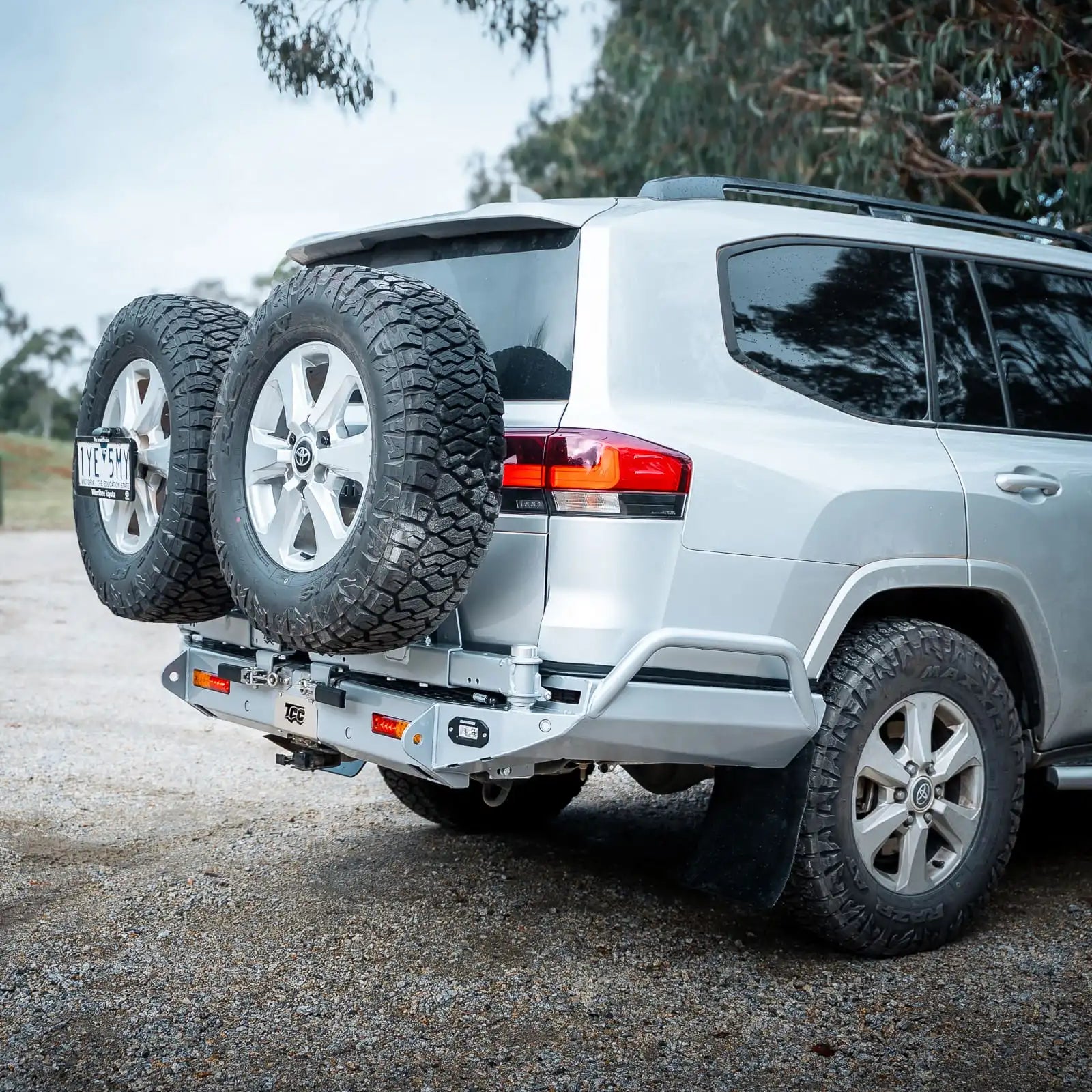 Silver SUV with a spare tire on a hitch, parked outdoors with trees in the background.