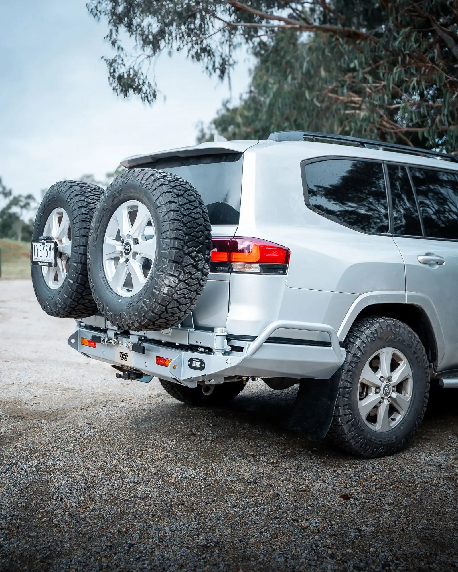 Silver SUV with a spare tire on a hitch, parked outdoors with trees in the background.