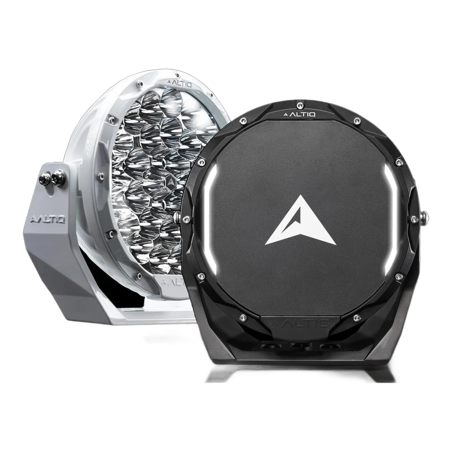 A white and a black ALTIQ Rogue 8.5 driving light photographed against black, the white one showing its reflector face and the black one its arrow-marked cover with the illuminated position-light strip curving across it