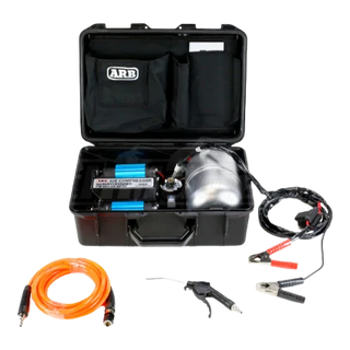 ARB twin piston compressor in its black travel case with the lid open, the twin blue cylinders and polished air tank inside, with the orange hose, blower gun and battery clamp leads laid out in front
