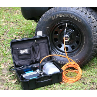 ARB twin piston compressor in its open travel case beside a vehicle on grass, the orange air hose connected to a Pro Comp wheel on a mud terrain tyre