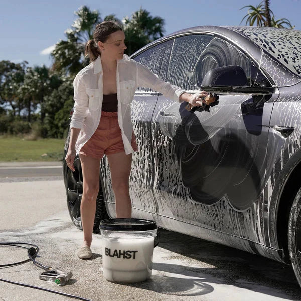 Woman washing a car with a sponge, using a bucket labeled 'Blahst' on a sunny day.