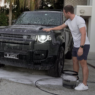Man washing a black Land Rover with a sponge and a bucket labeled 'BLAHST'.
